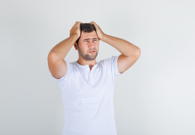 Young male holding head in hands in white t-shirt and looking negative