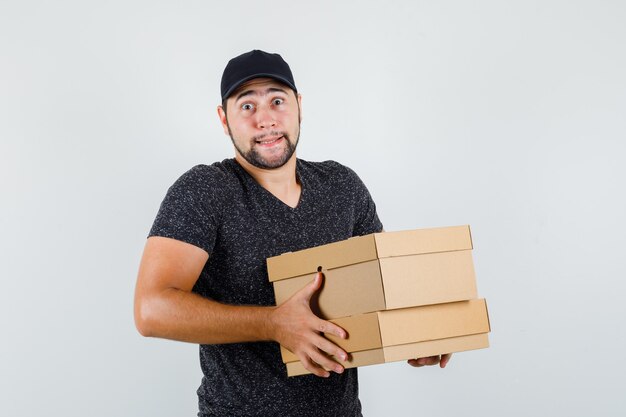 Young male holding heavy cardboard boxes in t-shirt and cap front view.