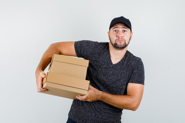 Young male holding heavy cardboard boxes in t-shirt and cap front view.