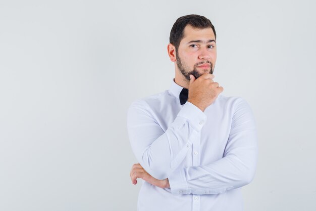 Young male holding his chin in white shirt and looking optimistic , front view.