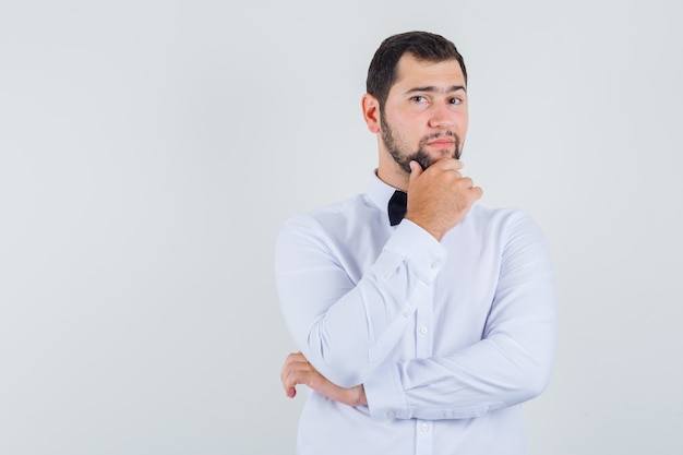 Young male holding his chin in white shirt and looking optimistic , front view.