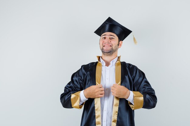 Young male holding his gown in graduate uniform and looking proud , front view.