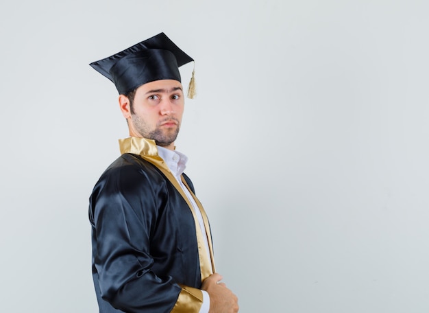 Young male holding his gown in graduate uniform and looking serious. front view.