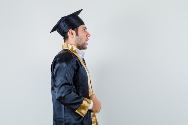 Молодой мужчина держит его платье в униформе выпускника и выглядит удивленным.