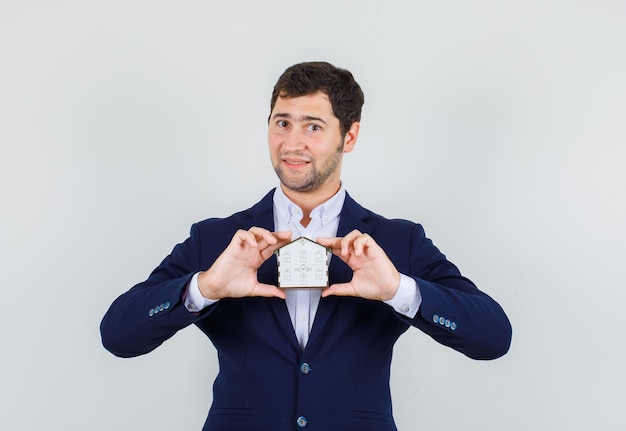 Young male holding house model in suit and looking cheerful. front view.