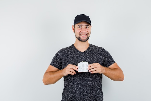 Young male holding house model in t-shirt and cap and looking cheerful