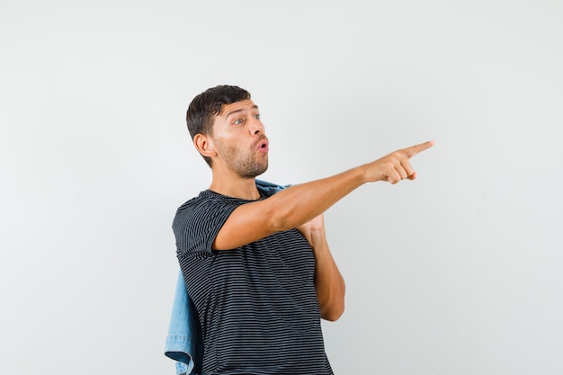 Young male holding jacket on back pointing away in t-shirt and looking surprised  