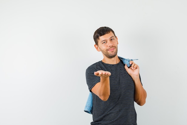 Young male holding jacket on back stretching palm in t-shirt and looking positive 