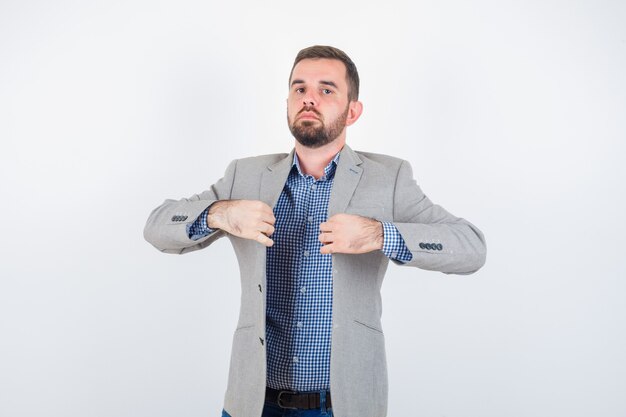 Free photo young male holding lapels while posing in shirt, jeans, suit jacket and looking serious , front view.