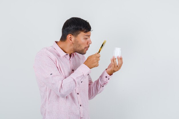 Young male holding magnifier over pill bottle in pink shirt front view.