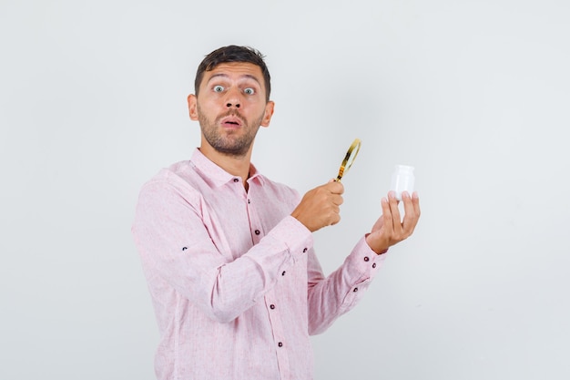 Young male holding magnifier over pill bottle in shirt and looking surprised , front view.