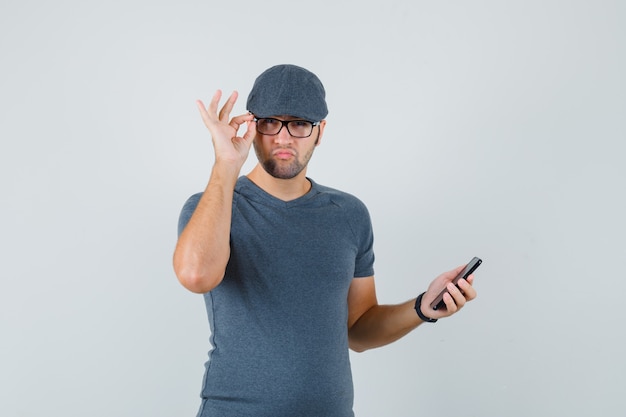 Молодой мужчина держит мобильный телефон в серой кепке футболки и выглядит сомнительно