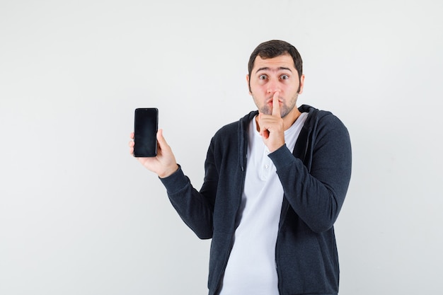 Молодой мужчина держит мобильный телефон, показывает жест молчания в футболке, куртке и выглядит обеспокоенным, вид спереди.