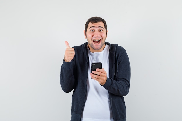 Молодой мужчина держит мобильный телефон, показывает палец вверх в футболке, куртке и рад, вид спереди.
