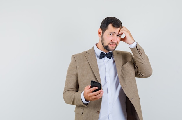 Young male holding mobile phone in suit and looking forgetful. front view.