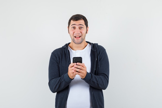 Young male holding mobile phone in t-shirt, jacket and looking glad. front view.