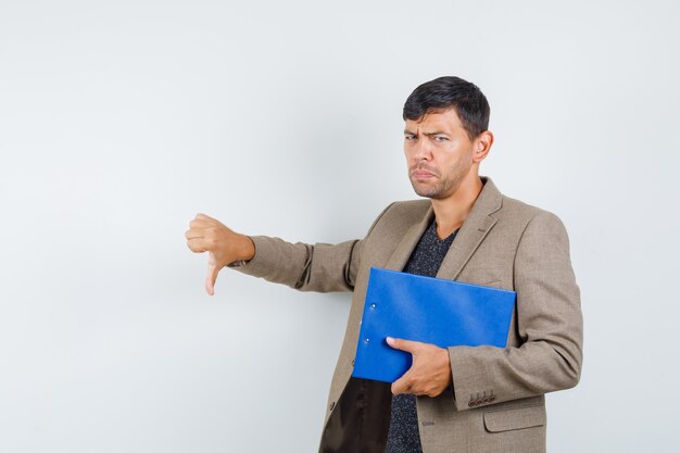 Young male holding notebook while showing thumb down in grayish brown jacket,black shirt and looking displeased. front view.