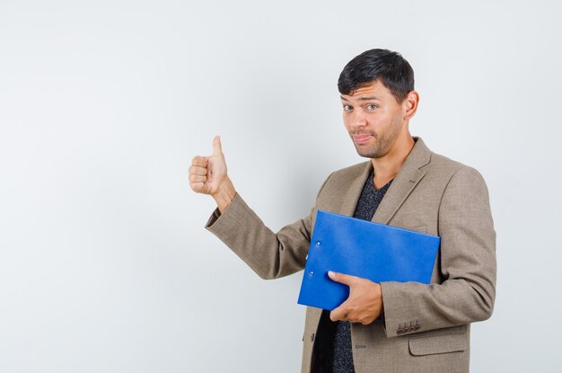 Молодой мужчина держит ноутбук, показывая большой палец вверх в серовато-коричневой куртке, черной рубашке и выглядит благодарным, вид спереди.