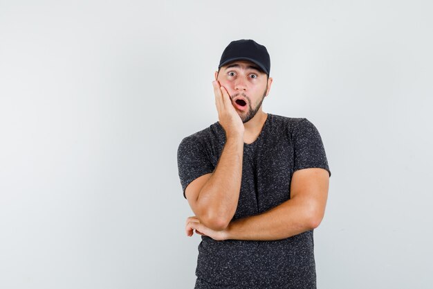 Young male holding palm on cheek in t-shirt and cap and looking surprised