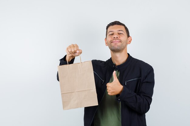 Молодой мужчина держит бумажный пакет с большим пальцем вверх в футболке, куртке и выглядит веселым. передний план.