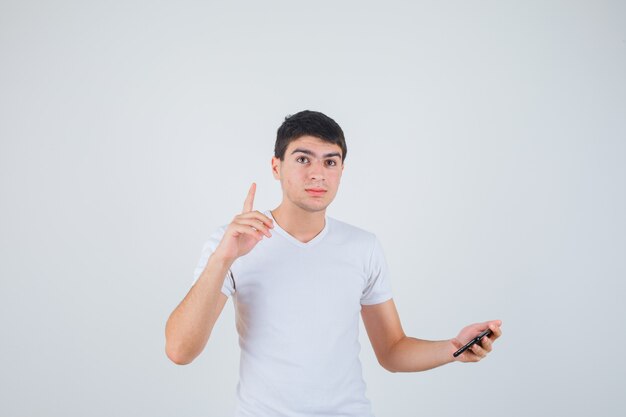 Young male holding phone, pointing up in t-shirt and looking self-confident , front view.