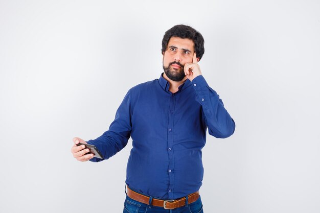 Young male holding phone while thinking in royal blue shirt , front view.