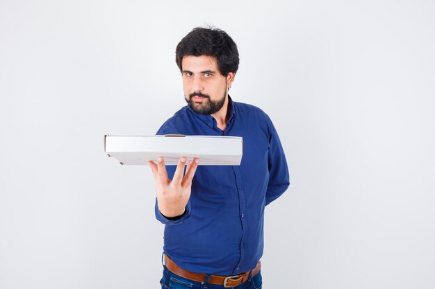 Young male holding pizza box in shirt, jeans and looking confident. front view.