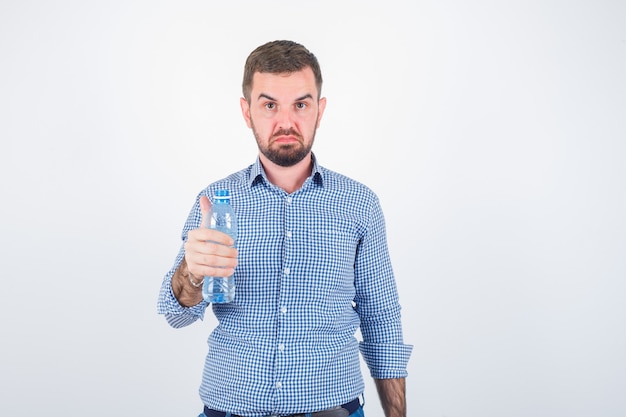 Young male holding plastic water bottle in shirt, jeans and looking serious. front view.