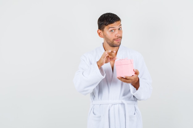 Young male holding present box in white bathrobe and looking curious. front view.