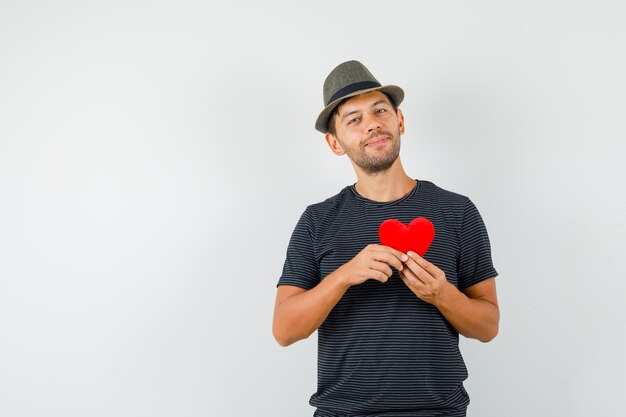 Young male holding red heart in t-shirt hat and looking cheerful 