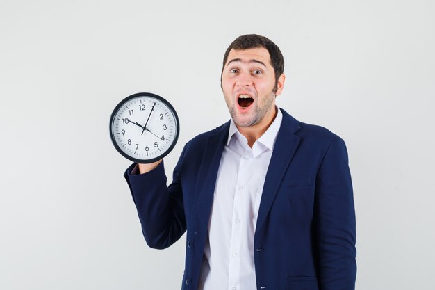 Young male holding wall clock in shirt and jacket and looking amazed