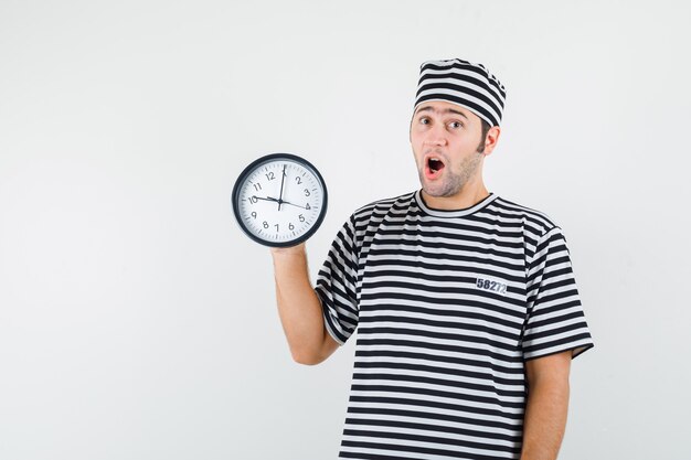 Young male holding wall clock in t-shirt, hat and looking amazed , front view.