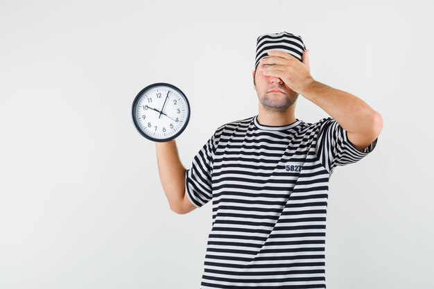 Young male holding wall clock in t-shirt, hat and looking forgetful , front view.