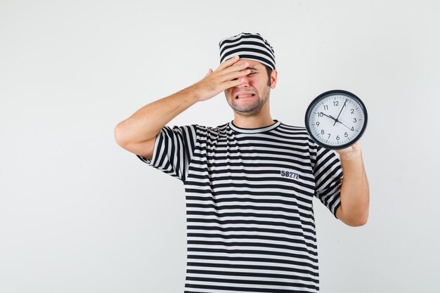 Young male holding wall clock in t-shirt, hat and looking mournful. front view.