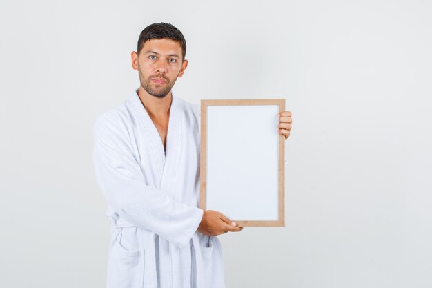 Young male holding white board in white bathrobe and looking strict , front view.