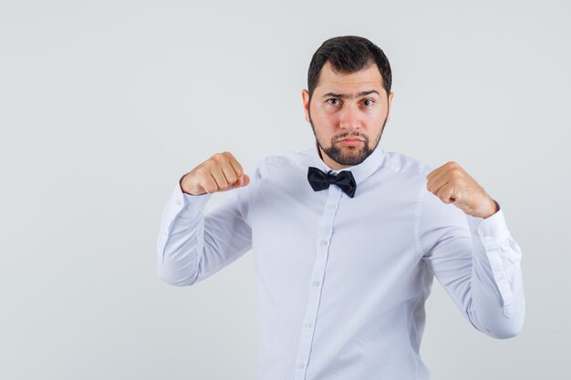 Young male keeping clenched fists raised in white shirt and looking serious. front view.