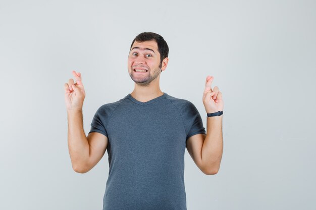 Young male keeping fingers crossed in grey t-shirt and looking cheerful 