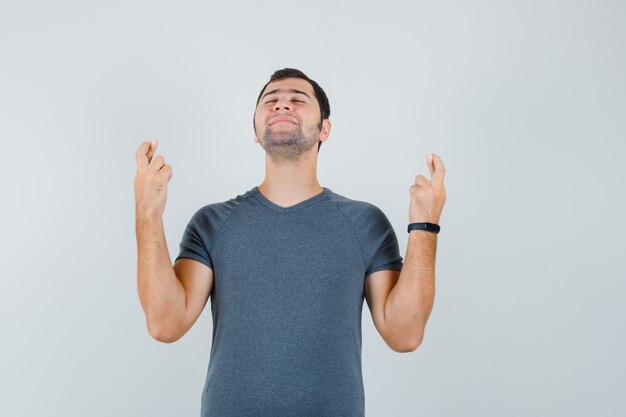 Young male keeping fingers crossed in grey t-shirt and looking peaceful  