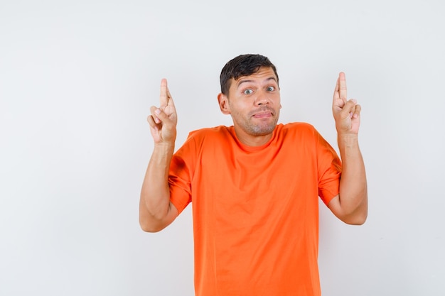 Young male keeping fingers crossed in orange t-shirt and looking optimistic