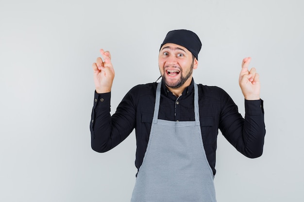 Young male keeping fingers crossed in shirt, apron and looking happy. front view.