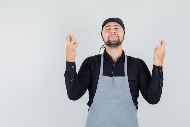 Young male keeping fingers crossed in shirt, apron and looking hopeful , front view.