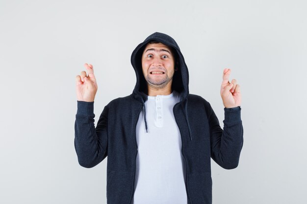 Young male keeping fingers crossed in t-shirt, jacket and looking cheery. front view.