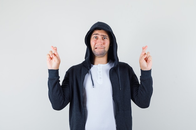 Young male keeping fingers crossed in t-shirt, jacket and looking troubled. front view.