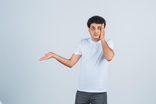 Young male keeping hand on cheek, showing something in white t-shirt, pants and looking puzzled. front view.