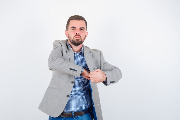 Young male keeping hand in pocket of jacket in shirt, jeans, suit jacket and looking confident. front view.