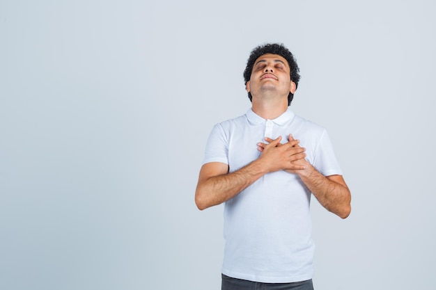 Young male keeping hands on chest in white t-shirt, pants and looking happy , front view.