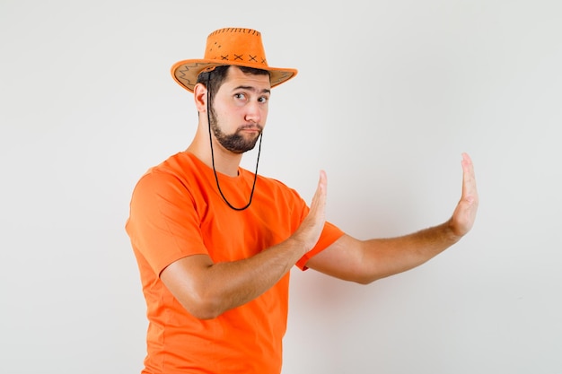 Молодой мужчина держит руки в защитной манере в оранжевой футболке, шляпе и смотрит внимательно, вид спереди.