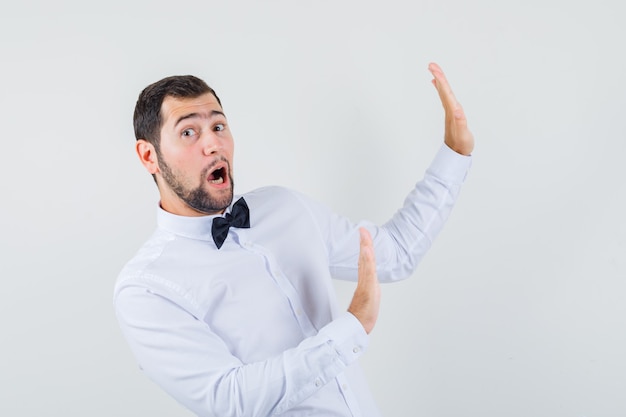 Young male keeping hands in protective manner in white shirt and looking scared. front view.