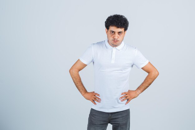 Young male keeping hands on waist in white t-shirt, pants and looking spiteful , front view.