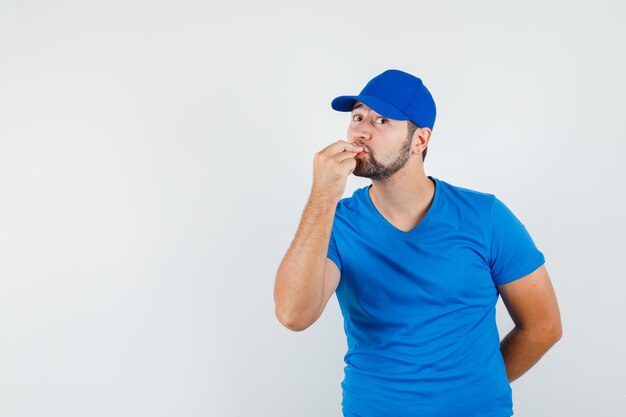 Young male kissing fingers while making delicious gesture in blue t-shirt and cap and looking joyful